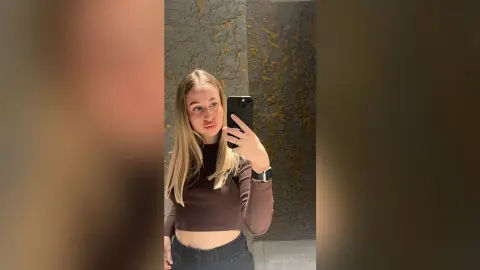 AnnelleToulson takes a mirror selfie wearing a brown long-sleeve crop top and black pants, showing her long blonde hair and face.