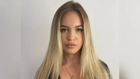 Blonde woman with long hair wearing a black top and silver necklace against a plain white background.