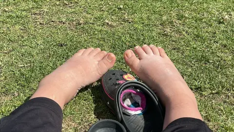 Bare feet resting on green grass next to black Crocs in a relaxing outdoor summer setting.