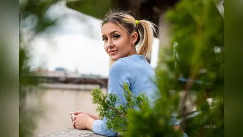 Anita looks back over her shoulder, wearing a light blue long-sleeved top with her blonde hair tied in a high ponytail.