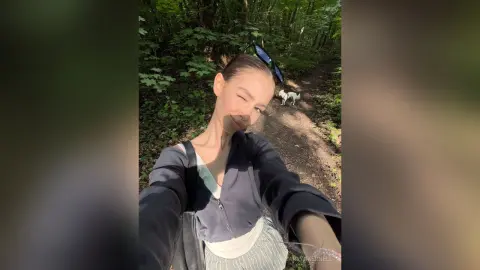 CarlynKernell wearing a dark zip-up jacket and light skirt, posing for a selfie on a sunlit forest path with a small white dog behind her.