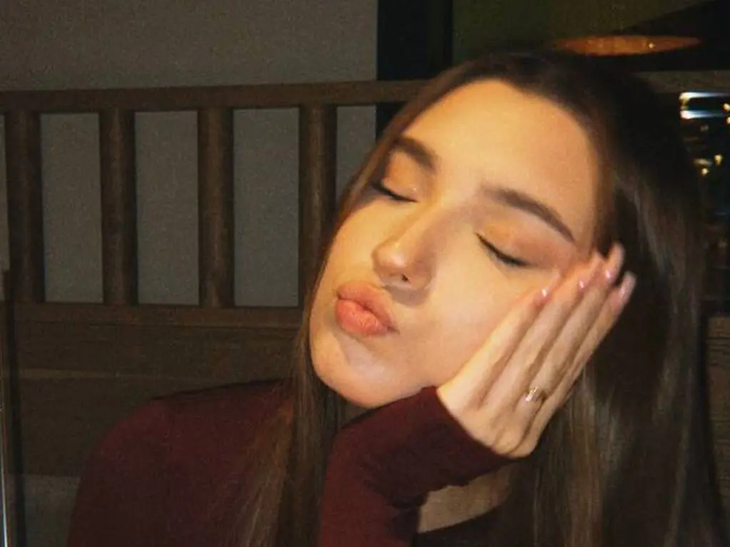 Young woman in a burgundy sweater making a playful pouty face in a dimly lit indoor setting.