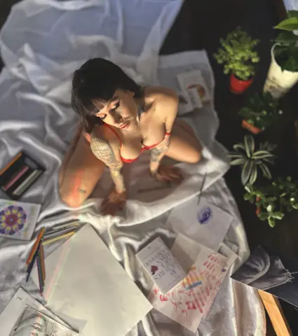 Young woman in red lingerie sitting on a white blanket surrounded by art supplies and houseplants in a cozy room.
