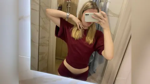 Blonde woman in a burgundy t-shirt taking a casual mirror selfie in a modern marble bathroom.