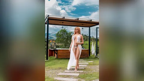 Blonde woman in a white lace cover-up standing on a stone path under a wooden pergola in a scenic mountain landscape.