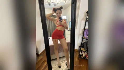 Young woman in a red mini dress taking a mirror selfie in a modern bedroom setting.