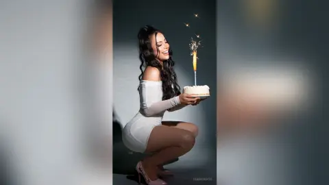 Smiling woman in a white dress holding a birthday cake with a sparkling candle against a dark studio background.