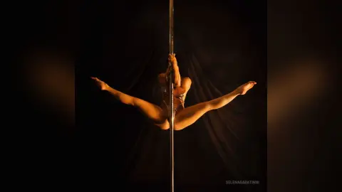 Athletic woman in a sparkly bodysuit performing a graceful split on a vertical pole against a dark backdrop.