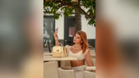 Young woman in a white summer outfit taking a selfie outdoors at a sunny cafe terrace.