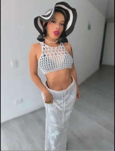 Young woman in white crochet crop top and matching skirt wearing a large black and white sun hat indoors.