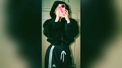 Young woman in a cozy black fuzzy hoodie and Adidas track pants taking a mirror selfie in a casual mood.