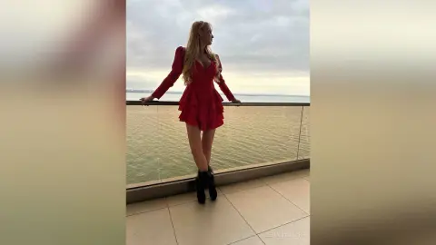 Blonde woman in a stylish red mini dress posing on a balcony overlooking the ocean at sunset.