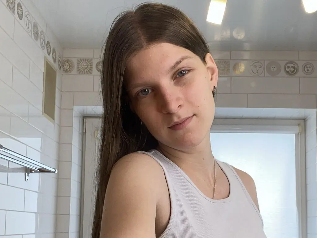 Young woman with long brown hair wearing a white ribbed tank top in a bright, modern tiled bathroom.