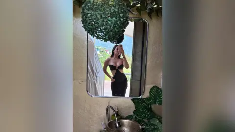 Stylish woman in a black cutout maxi dress taking a mirror selfie in a bright, modern tropical room.
