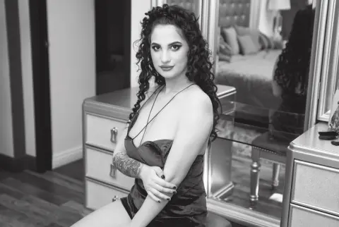Glamorous woman with curly hair in a dark satin slip dress posing in a modern bedroom setting.