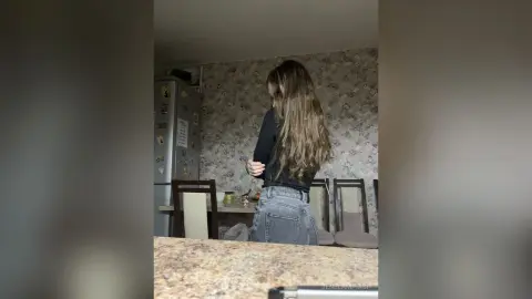 Young woman with long hair wearing a black long-sleeve shirt and denim jeans in a floral patterned room.
