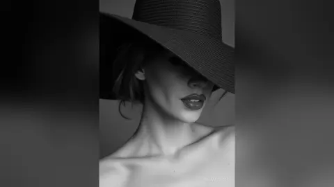 AnaMathers poses head-on wearing a wide-brimmed black hat and dark lipstick, showing her bare shoulders and neck.