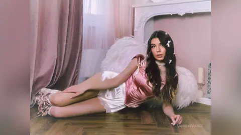 Young woman in white angel wings and pink satin corset relaxing on a wooden floor in a dreamy bedroom setting.