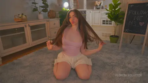 Young woman in pink tank top and lace shorts sitting on a rug in a cozy, bright living room.