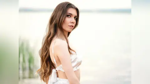 Young woman with long wavy hair in a white summer dress posing by a serene lake at sunset.