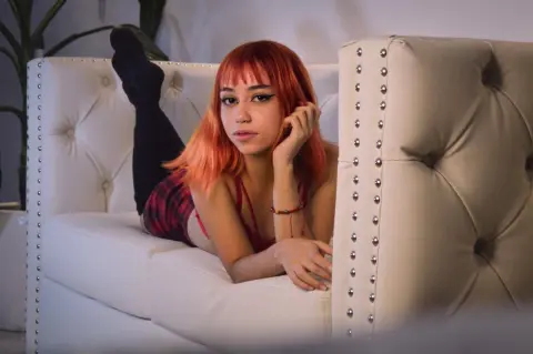 Young woman with red hair in a red plaid outfit relaxing on a white tufted sofa in a cozy indoor setting.