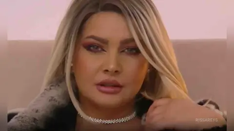 Glamorous blonde woman with dramatic eye makeup and silver choker posing in a soft-lit indoor setting.