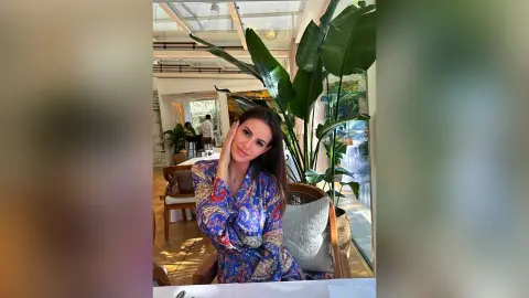 Woman in a blue floral patterned dress sitting in a bright sunlit indoor garden restaurant.