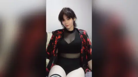 Young woman in a black lace bodysuit and floral kimono sitting in a minimalist indoor setting.