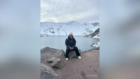 Woman in black winter jacket and pink beanie sitting on rocks by a snowy mountain lake under a cloudy sky.