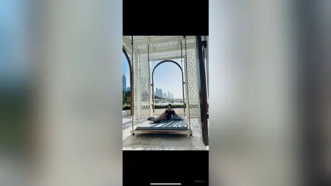 Woman in black outfit relaxing on a large swing bed overlooking a modern city skyline at a luxury resort.