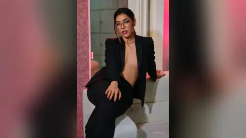 JessieLiu wears a black blazer and matching trousers while sitting on a white sofa, looking at the camera with glasses on.