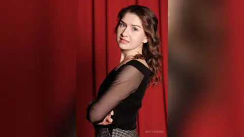 Smiling young woman in a black sheer blouse posing against a red velvet curtain background.