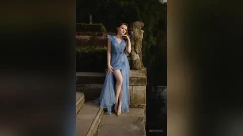 RubySoul poses in a long, light blue dress with a high slit, standing outdoors near a stone wall and a statue.