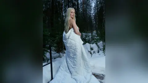 EllieFrosty stands in a snowy forest, wrapped in a thick white duvet that drapes over her body and exposes her bare shoulders and chest.