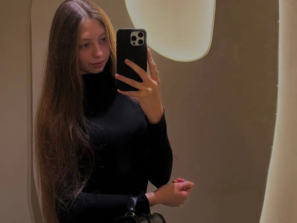 Young woman with long hair in a black turtleneck taking a moody mirror selfie in warm indoor lighting.