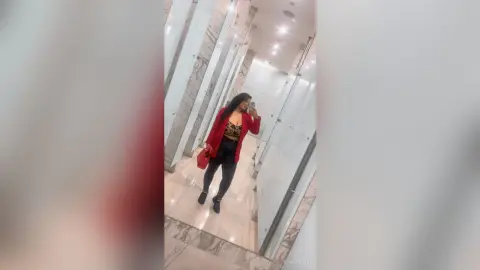 NiaCorelli poses in a red blazer and black leggings, holding a red bag in a bright marble hallway.