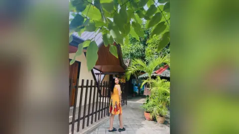 Cheerful woman in a yellow floral sundress standing by a wooden fence in a lush tropical garden setting.