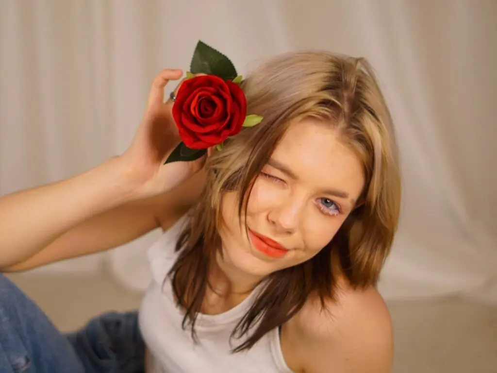 Blonde woman in a white tank top holding a red rose near her face in a soft, dreamy studio setting.