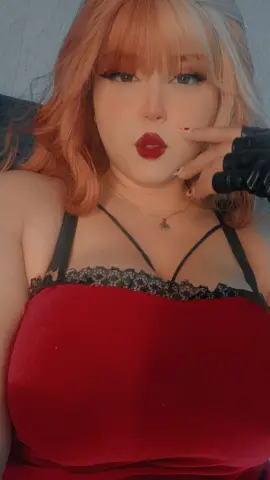 Woman with wavy ginger hair wearing a red velvet dress and black lace trim in a soft indoor setting.