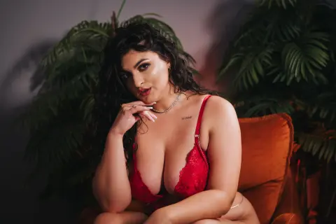 Curvy woman in red lace lingerie sitting in a moody indoor setting with lush green tropical plants.