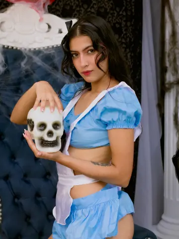 SusanaSanchez20 wears a light blue, short-sleeved crop top and matching skirt while holding a white skull prop against a dark, tufted chair background.