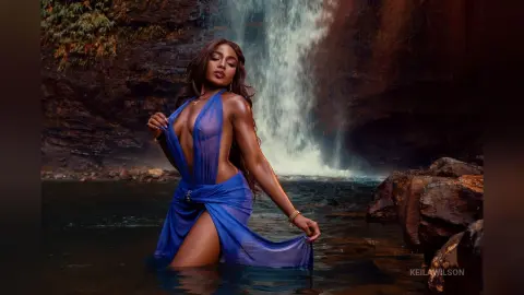 Elegant woman in a flowing blue dress posing by a misty waterfall in a serene jungle setting.