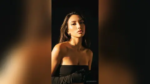 Elegant woman in a black strapless dress and gloves posing under warm cinematic lighting against a dark background.