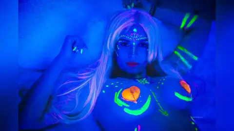 AndreaKalo lies on a bed with long blonde hair and glowing neon green body paint decorating her face and breasts under blue UV light.