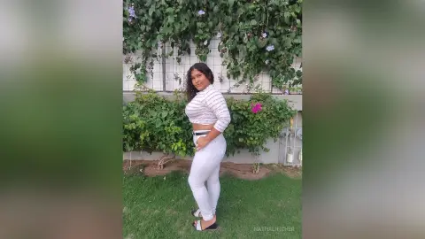 Smiling woman in a white striped crop top and white jeans posing in a lush green garden setting.