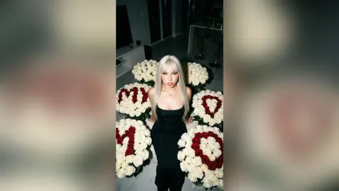 Elegant woman in a black evening gown sitting amidst large white rose arrangements in a modern room.