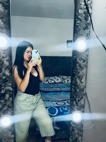 Young woman in a black tank top and light denim jeans taking a selfie in a lighted vanity mirror.