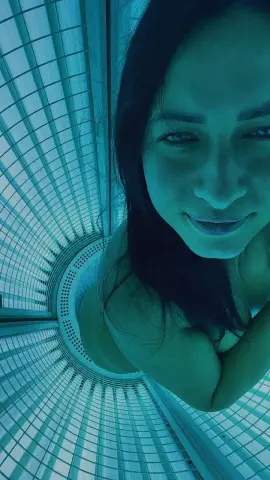 Smiling woman in a teal indoor setting with modern architectural window patterns and soft lighting.