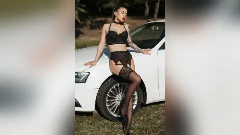 Glamorous woman in black lace lingerie and stockings posing by a white car in a sunny outdoor setting.