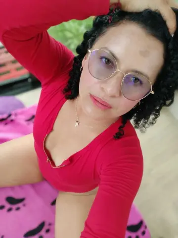 Smiling woman in a red long-sleeve top and glasses relaxing on a pink patterned surface.